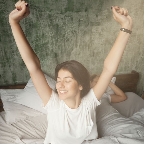A woman in a white shirt stretches and smiles in bed, eyes closed, with another person lying under the covers next to her against a green textured wall.